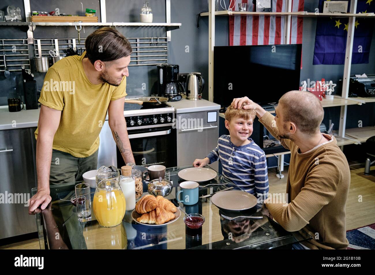 Feliz niño pequeño y dos hombres jóvenes hablando por mesa en la cocina Foto de stock