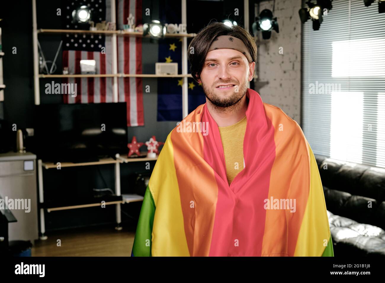 Hombre joven feliz envuelto en una bandera de orgullo de pie en el salón Foto de stock