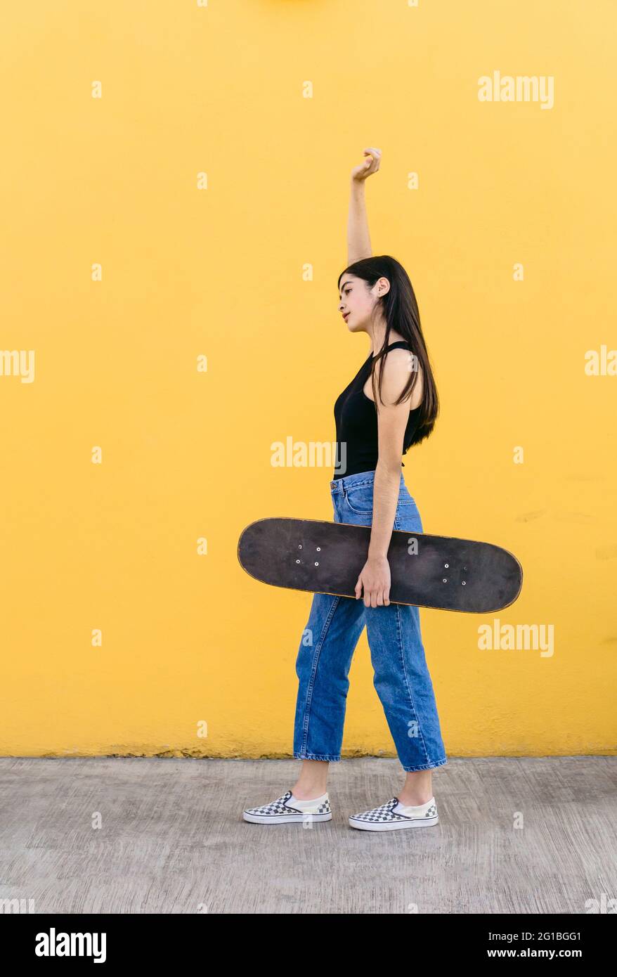 Vista lateral de la joven patinadora femenina con el brazo elevado y el skateboard de pie mirando hacia fuera en la pasarela con colorida pared amarilla en el fondo en daytim Foto de stock