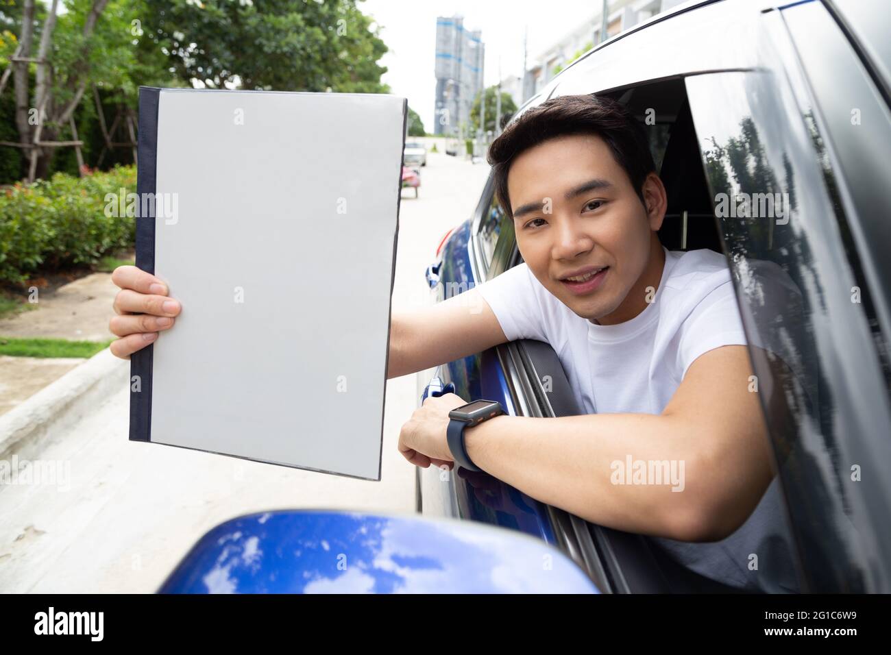 Hombre asiático joven que sostiene el libro de la póliza del coche y que se sienta en el coche, concepto del seguro personal del accidente Foto de stock