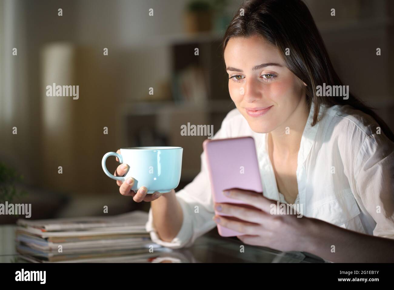 Mujer satisfecha sosteniendo una taza de café comprobando el teléfono inteligente en la noche en casa Foto de stock