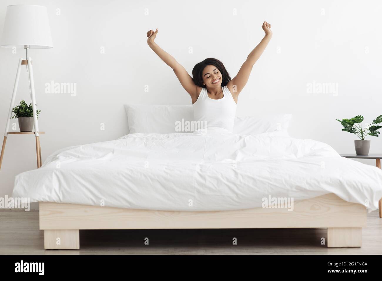 Feliz mujer afroamericana sentada en la cama, estirando los brazos después de dormir y disfrutando de la mañana en el acogedor dormitorio Foto de stock