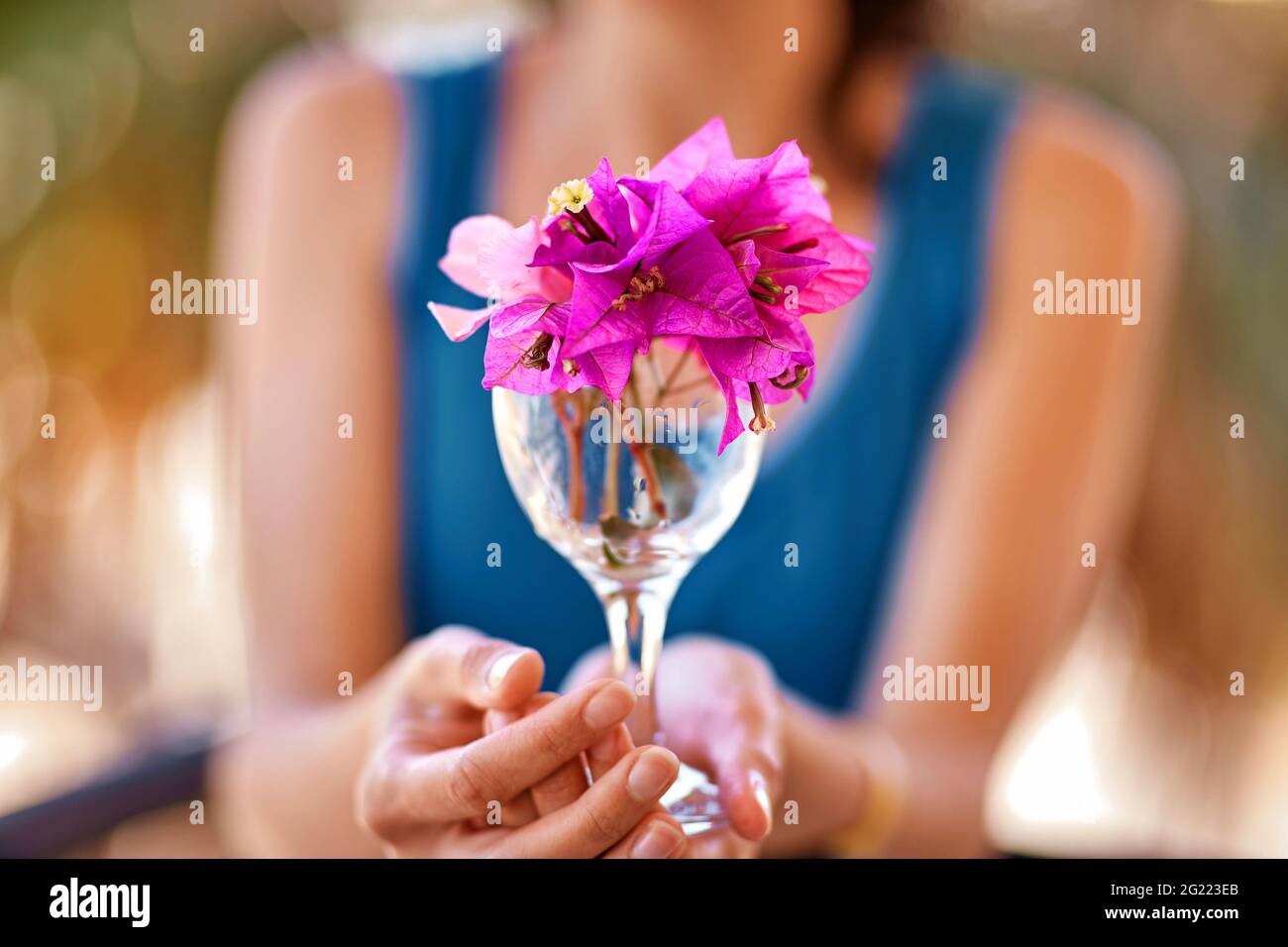 Mujer sostiene flores rosas brillantes Bougainvillea en un vaso de vino delante de sí misma. Concepto publicitario de viajes y vacaciones de verano. Verano flores surrealistas creativo concepto de moda Foto de stock
