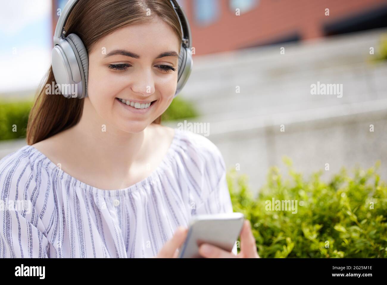 Sonriente joven estudiante en el campus universitario transmite música desde el teléfono móvil a los auriculares inalámbricos Foto de stock
