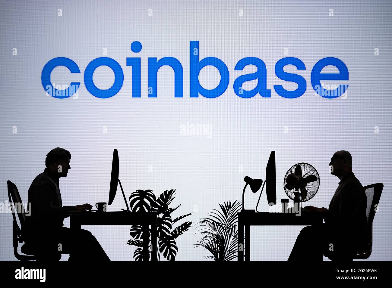 El logotipo de Coinbase se ve en una pantalla LED de fondo, mientras que dos personas con siluetas trabajan en un entorno de oficina (sólo para uso editorial) Foto de stock