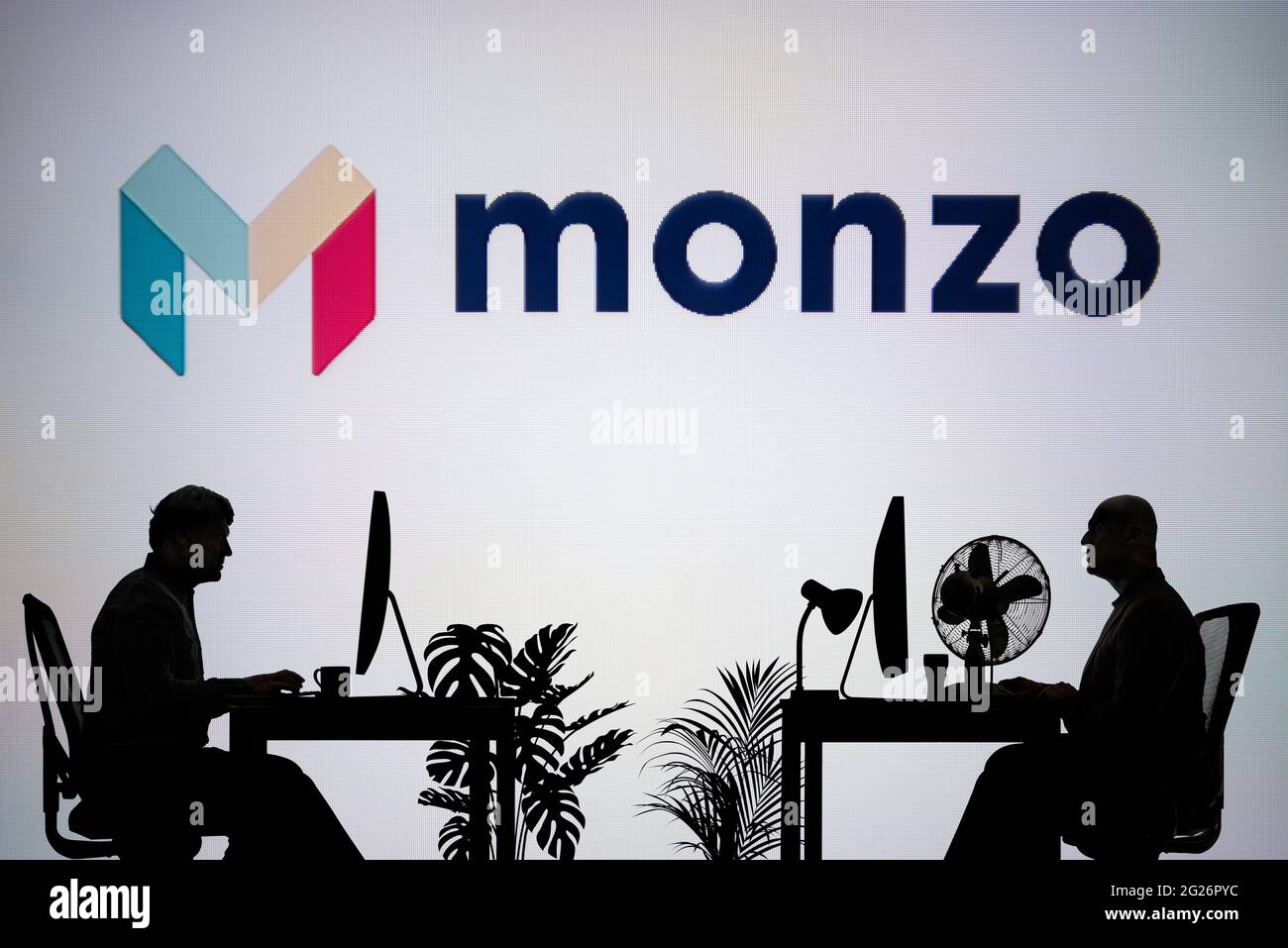 El logotipo del banco Monzo se ve en una pantalla LED de fondo, mientras que dos personas con siluetas trabajan en un entorno de oficina (sólo para uso editorial) Foto de stock