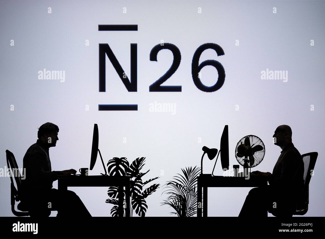 El logotipo del Banco N26 se ve en una pantalla LED en segundo plano, mientras que dos personas con siluetas trabajan en un entorno de oficina (sólo para uso editorial) Foto de stock