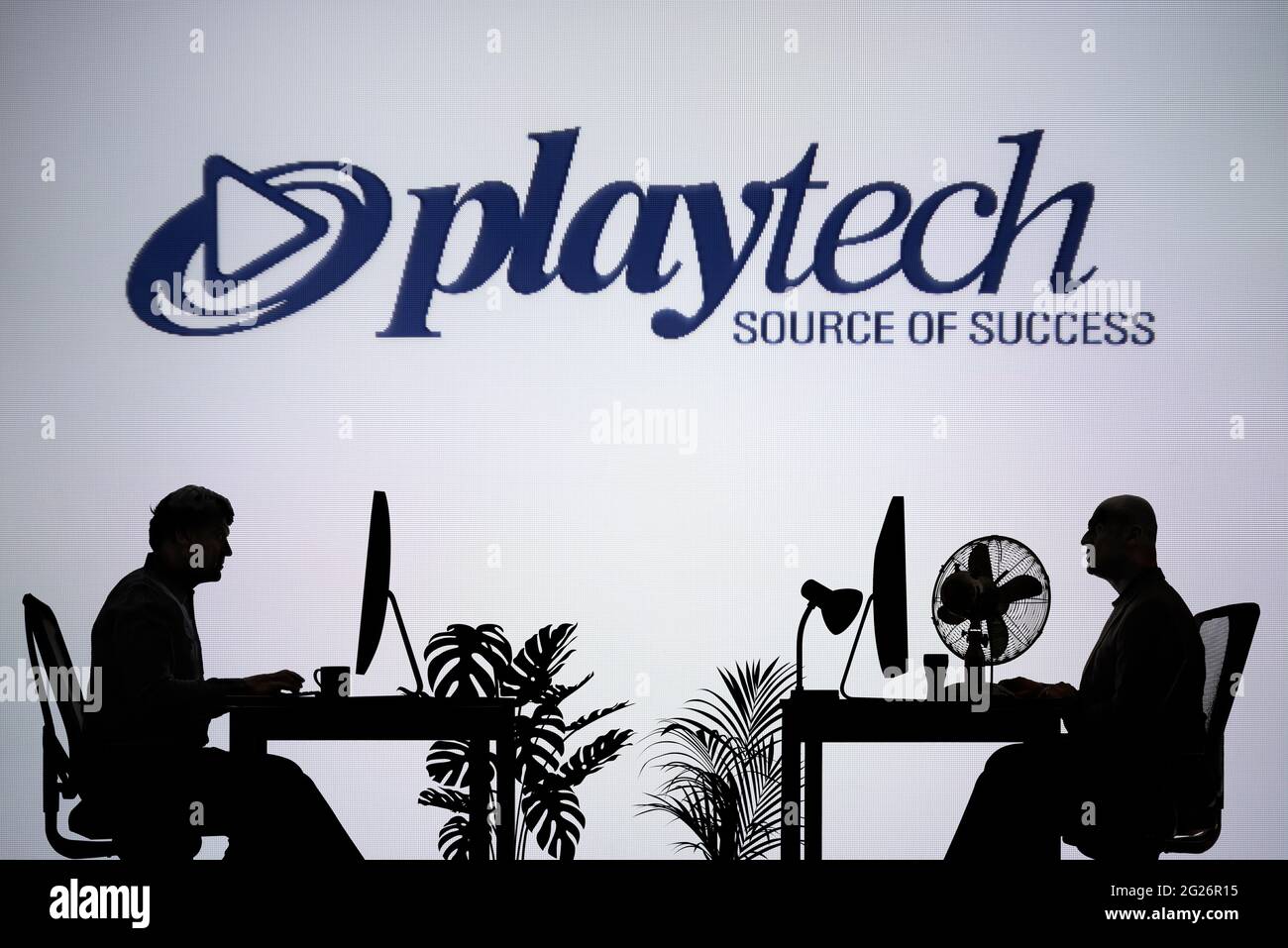 El logotipo de Playtech se ve en una pantalla LED de fondo, mientras que dos personas con siluetas trabajan en un entorno de oficina (sólo para uso editorial) Foto de stock