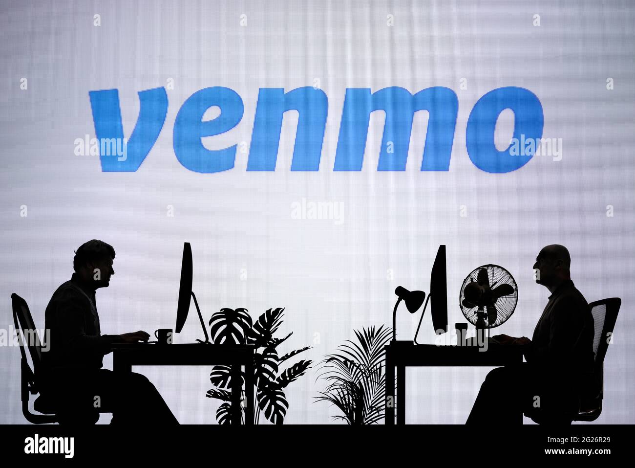 El logotipo de Venmo se ve en una pantalla LED de fondo, mientras que dos personas con siluetas trabajan en un entorno de oficina (sólo para uso editorial) Foto de stock