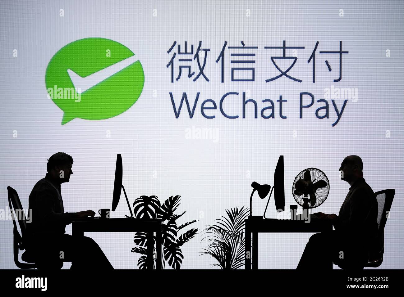 El logotipo de WeChat Pay se ve en una pantalla LED en segundo plano, mientras que dos personas con siluetas trabajan en un entorno de oficina (sólo para uso editorial) Foto de stock