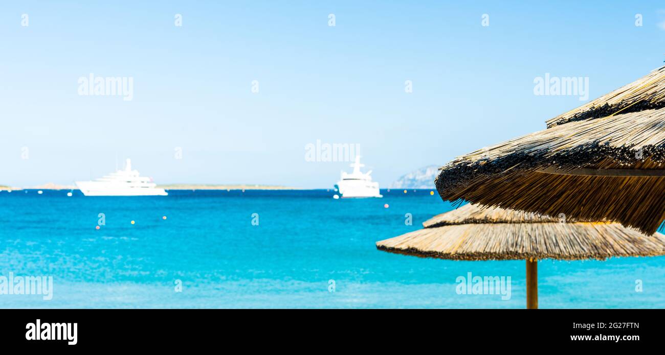 (Enfoque selectivo) Vista impresionante de una sombrilla en primer plano y dos yates de lujo navegando en un hermoso mar turquesa en el fondo. Foto de stock