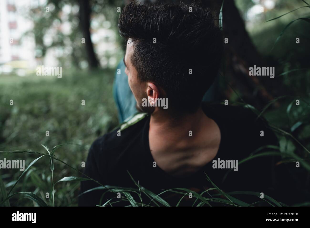 Hombre guapo en el bosque estira sus pies hasta un árbol y se encuentra en la vista trasera de hierba húmeda Foto de stock