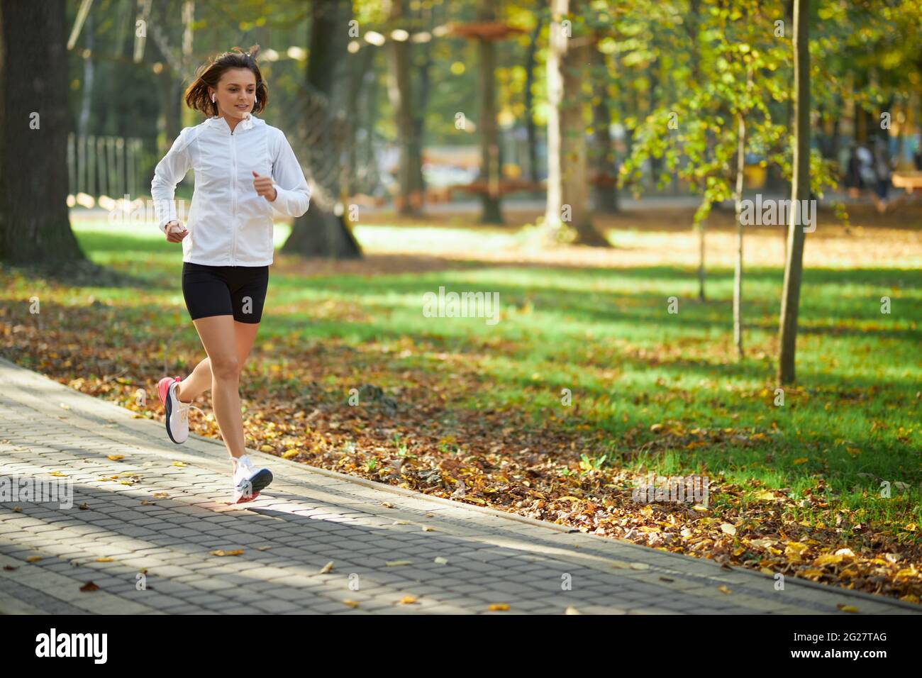 Mujer joven activa en ropa deportiva corriendo en el parque verde durante la mañana. Feliz mujer con el pelo oscuro disfrutando de la actividad deportiva en el aire fresco. Foto de stock