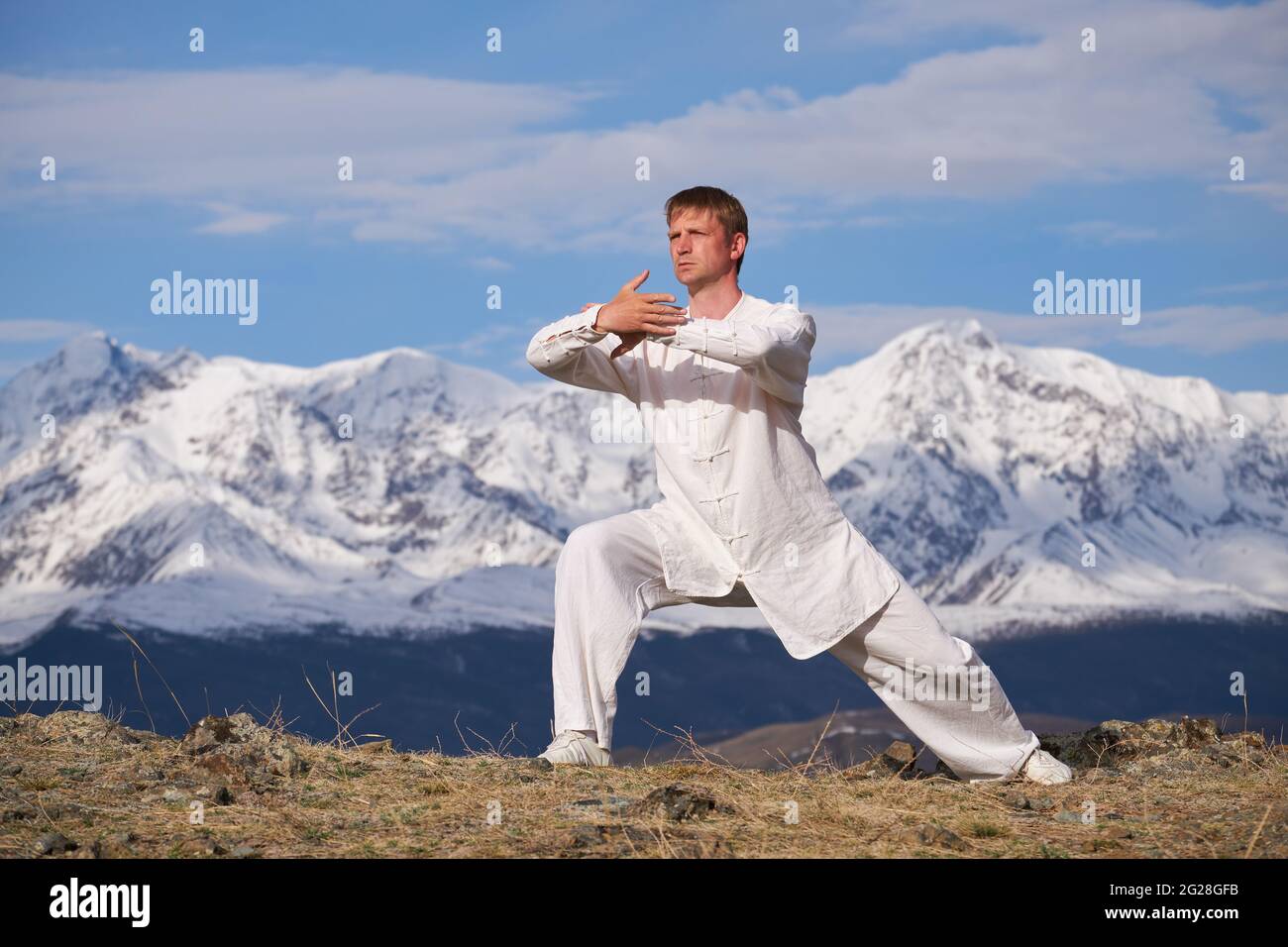 Wushu maestro en un entrenamiento de uniformes deportivos blancos en la colina. El campeón de Kungfu entrena las artes maritiales en la naturaleza sobre el fondo de las montañas nevadas. Foto de stock