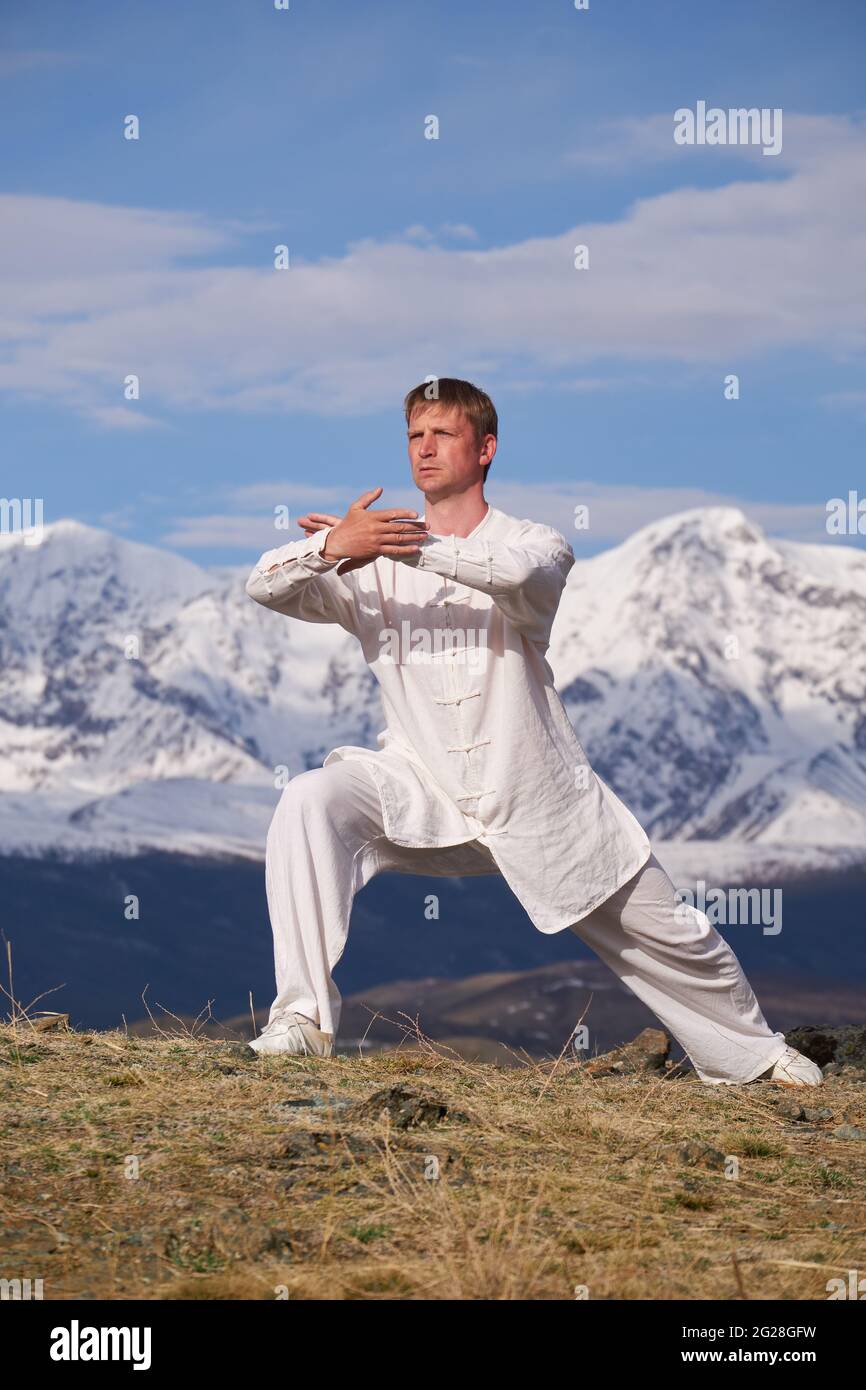 Wushu maestro en un entrenamiento de uniformes deportivos blancos en la colina. El campeón de Kungfu entrena las artes maritiales en la naturaleza sobre el fondo de las montañas nevadas. Foto de stock