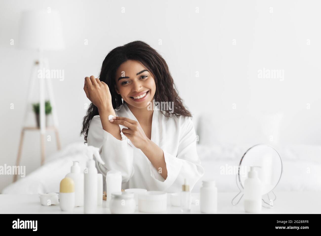 Cosméticos para el cuidado de la piel. Feliz mujer afroamericana aplicando crema de manos, hidratando la piel después del baño Foto de stock