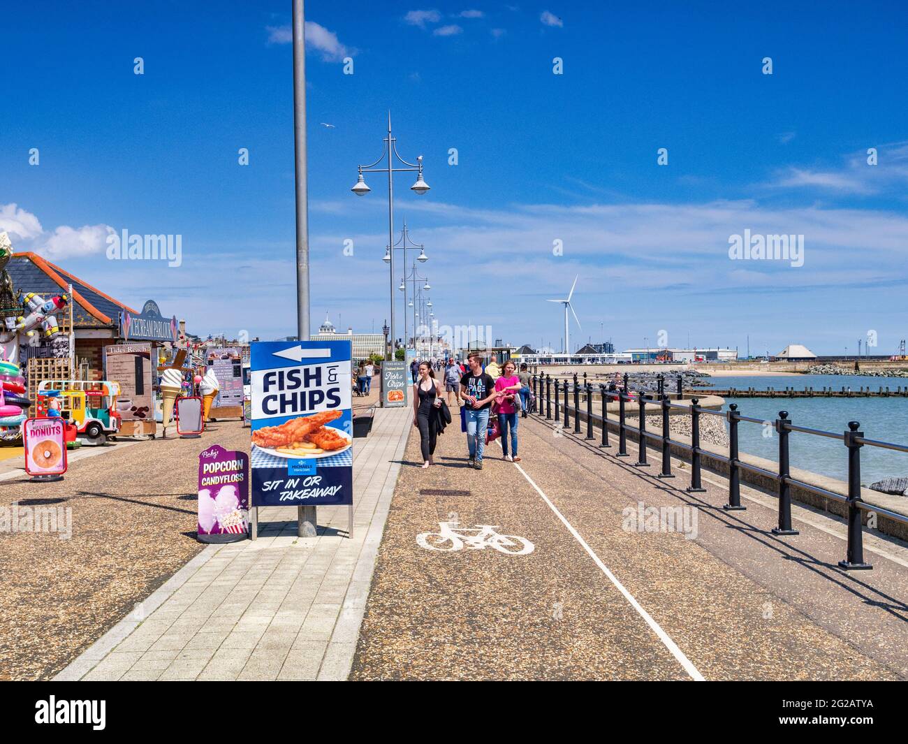 15 de junio de 2019: Lowestoft, Suffolk, Reino Unido - Pareja joven paseando por el paseo marítimo en un soleado día de verano en la costa, signo de pescado y patatas fritas. Foto de stock