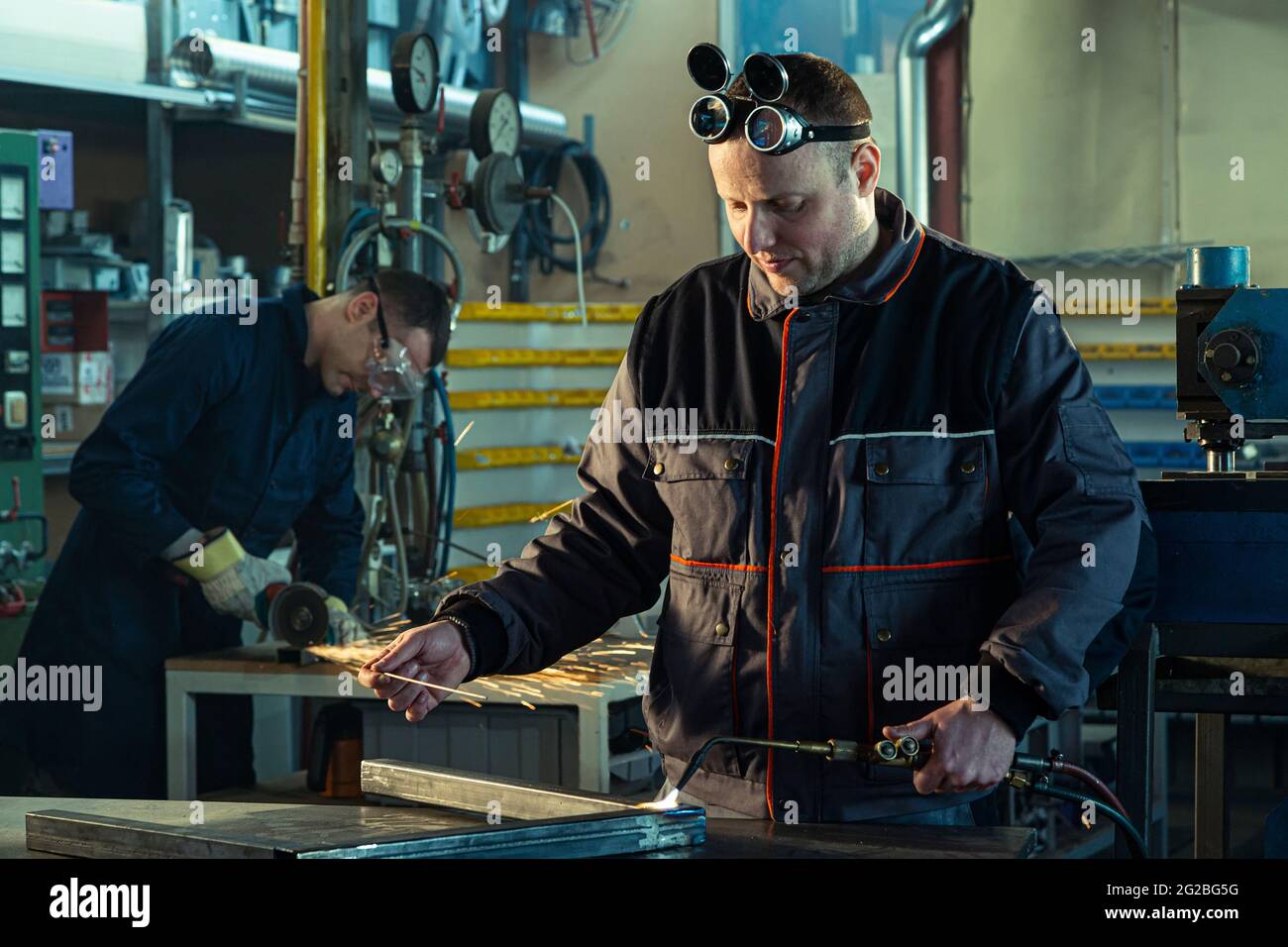 El soldador trabaja con una herramienta de soldadura autógena en el taller,  mientras que su compañero puede ser notado en el rectificado de espalda  Fotografía de stock - Alamy