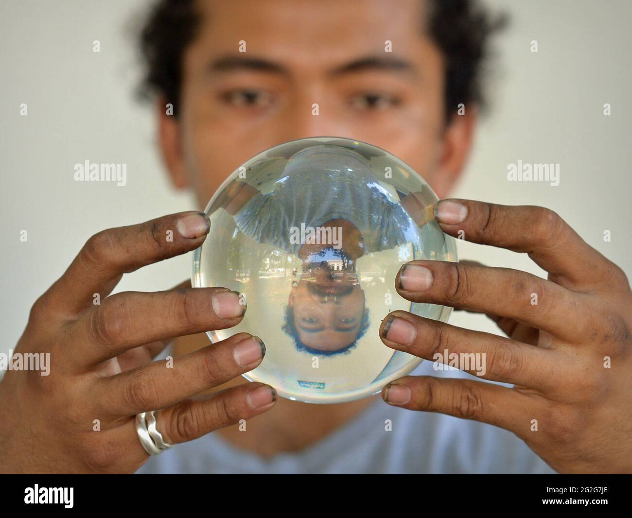 Un joven latino con uñas sucias sostiene una bola de cristal y mira fijamente con ojos fascinantes a su imagen de espejo volteada verticalmente en la orbe mágica. Foto de stock