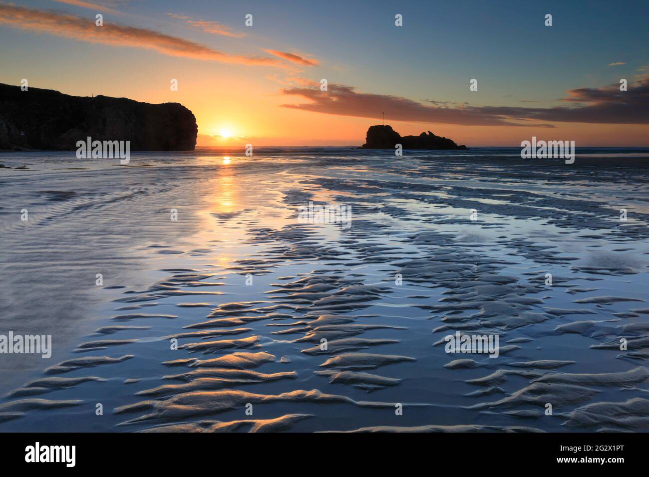 Ondulaciones de arena y un río lleva el ojo del espectador hacia Chapel Rock en la playa de Perranporth de Cornwall. La imagen se capturó poco antes de la puesta de sol Foto de stock