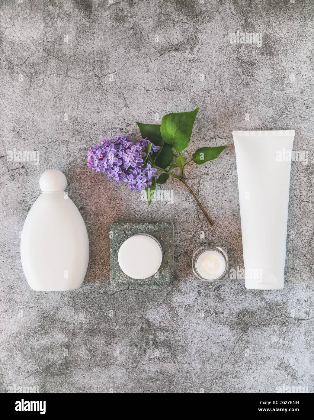 Composición natural del producto de belleza con botella, tubo y tarro de plástico blanco con ramita de lila sobre un fondo gris texturizado Foto de stock