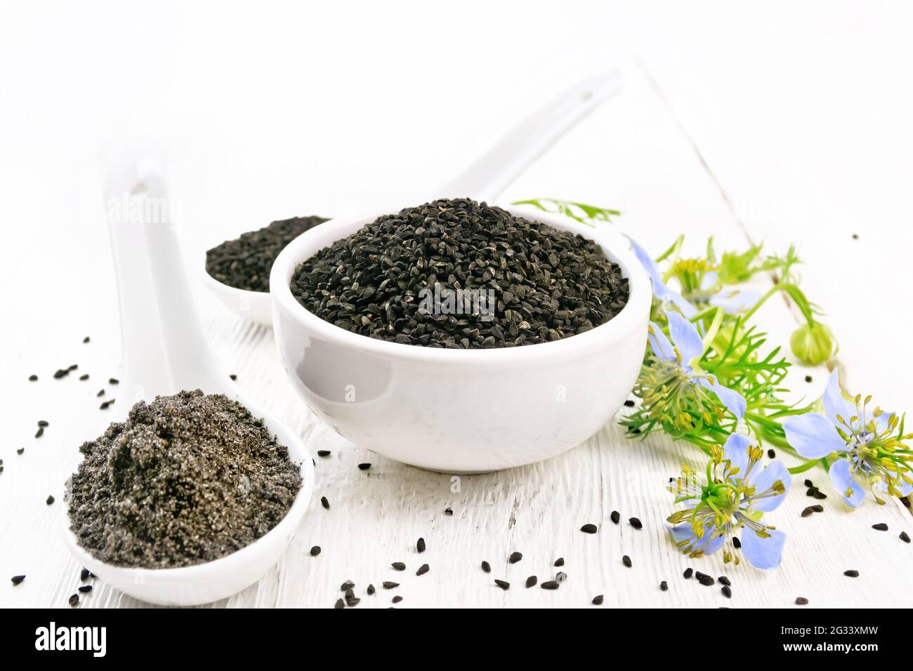 Semillas de comino negro en un bol, harina y semillas en cucharas, ramitas  de kalingi con flores azules y hojas verdes sobre el fondo de una tabla de  madera clara Fotografía de