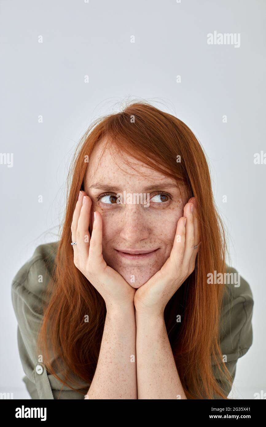 Mujer positiva con pelo de jengibre manteniendo las manos en las mejillas y mirando hacia fuera sobre fondo blanco Foto de stock