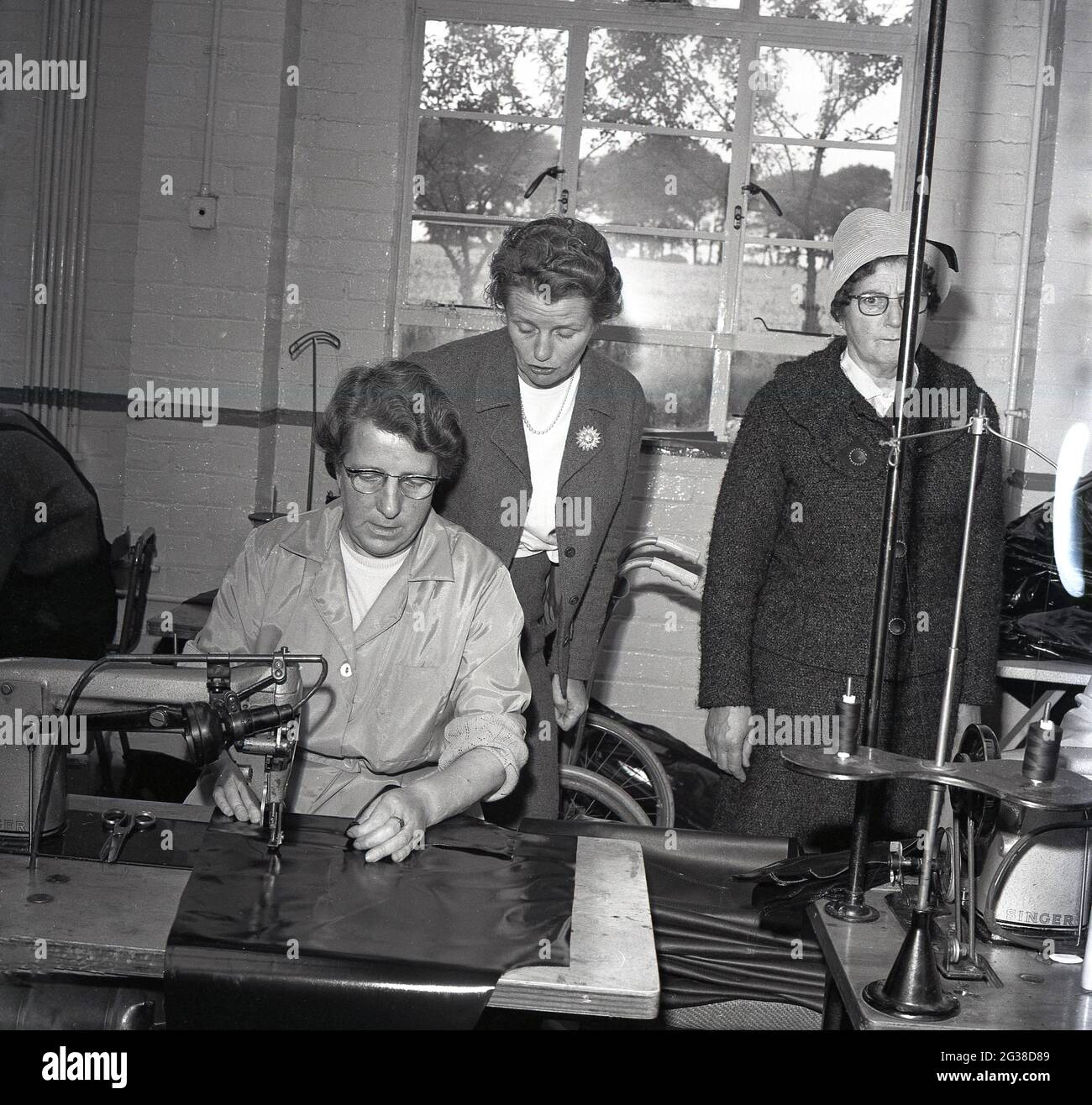1960s, histórico, dentro de una unidad industrial, dos concejales locales, de pie por una trabajadora, siendo re-empleado en la industria, utilizando una máquina para coser o sellar láminas protectoras, Cowdenbeath, Fife, Escocia. Foto de stock