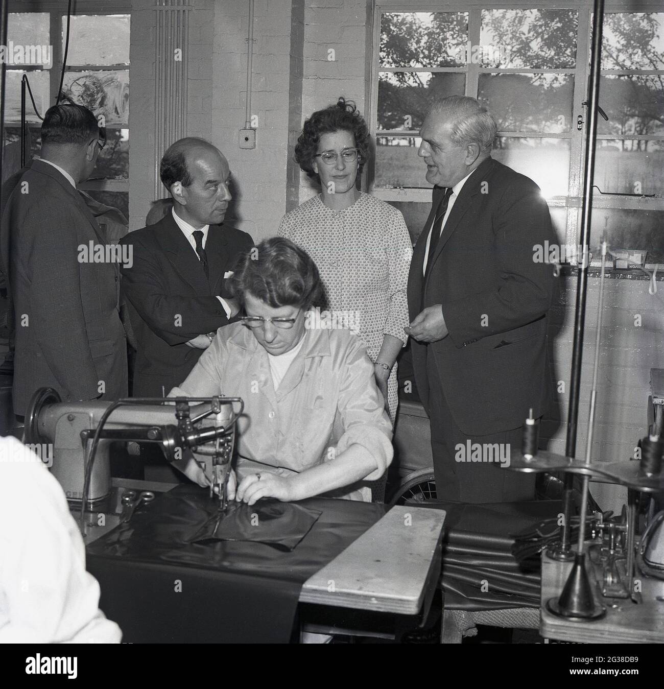 1960s, histórico, dentro de una unidad industrial, concejales locales y una empresa Directores, de pie por una trabajadora, siendo re-empleado en la industria, utilizando una máquina de coser de tipo industrial para coser hojas de cuero, Cowdenbeath, Fife, Escocia. Foto de stock