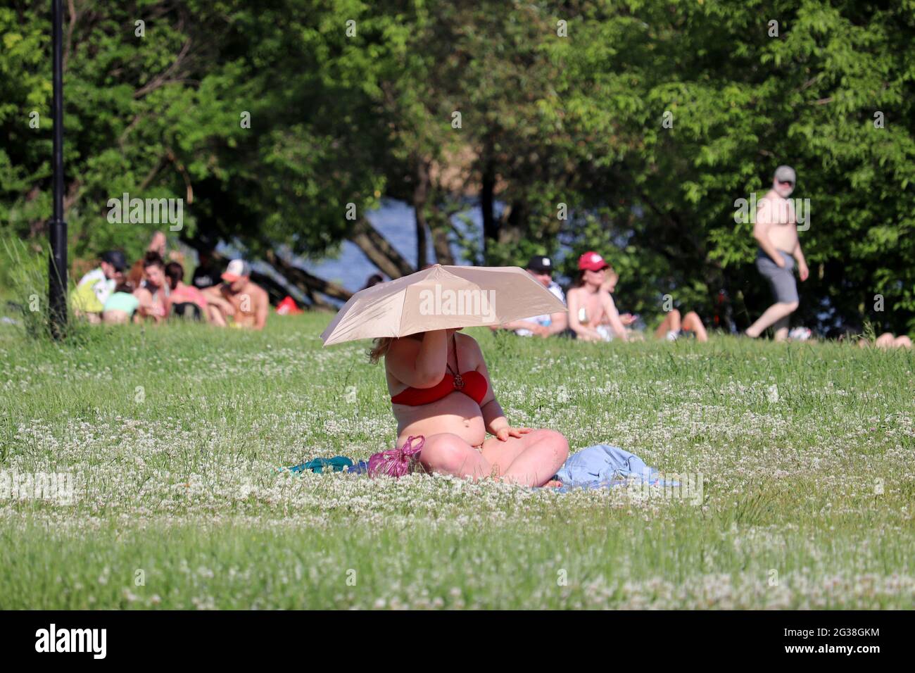 Mujer gorda en bikini bronceándose en una playa bajo el paraguas. Mujer sentada en una hierba en el parque de verano sobre fondo de la gente, fin de semana en una naturaleza Foto de stock