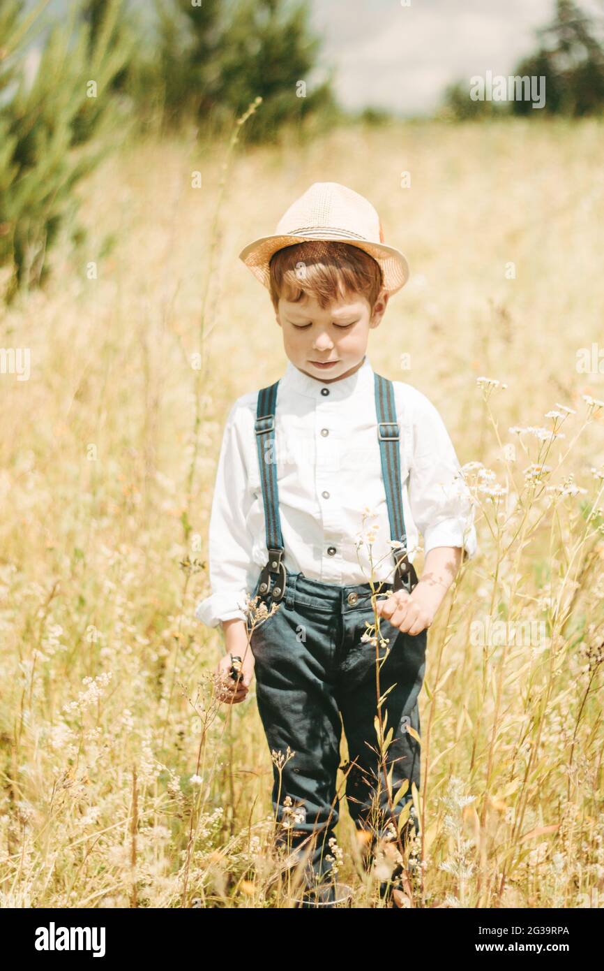 Un pequeño granjero camina en un campo de verano, un niño pequeño en un sombrero de paja. Niño en botas goma y una camisa blanca. Estilo campestre Fotografía de stock -