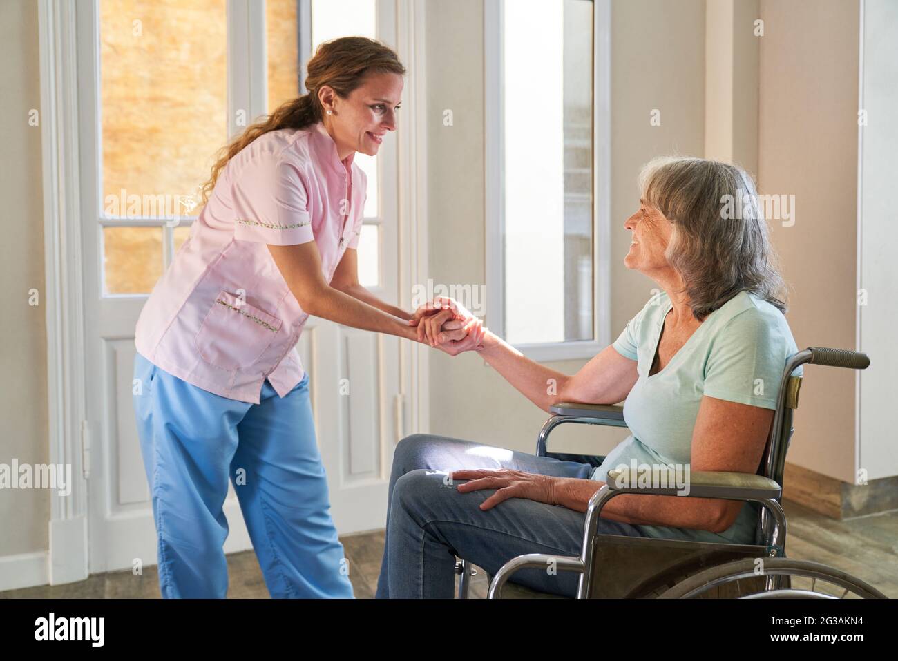 La mujer del servicio de enfermería mantiene la mano de un ciudadano mayor en silla de ruedas en el cuidado del hogar Foto de stock