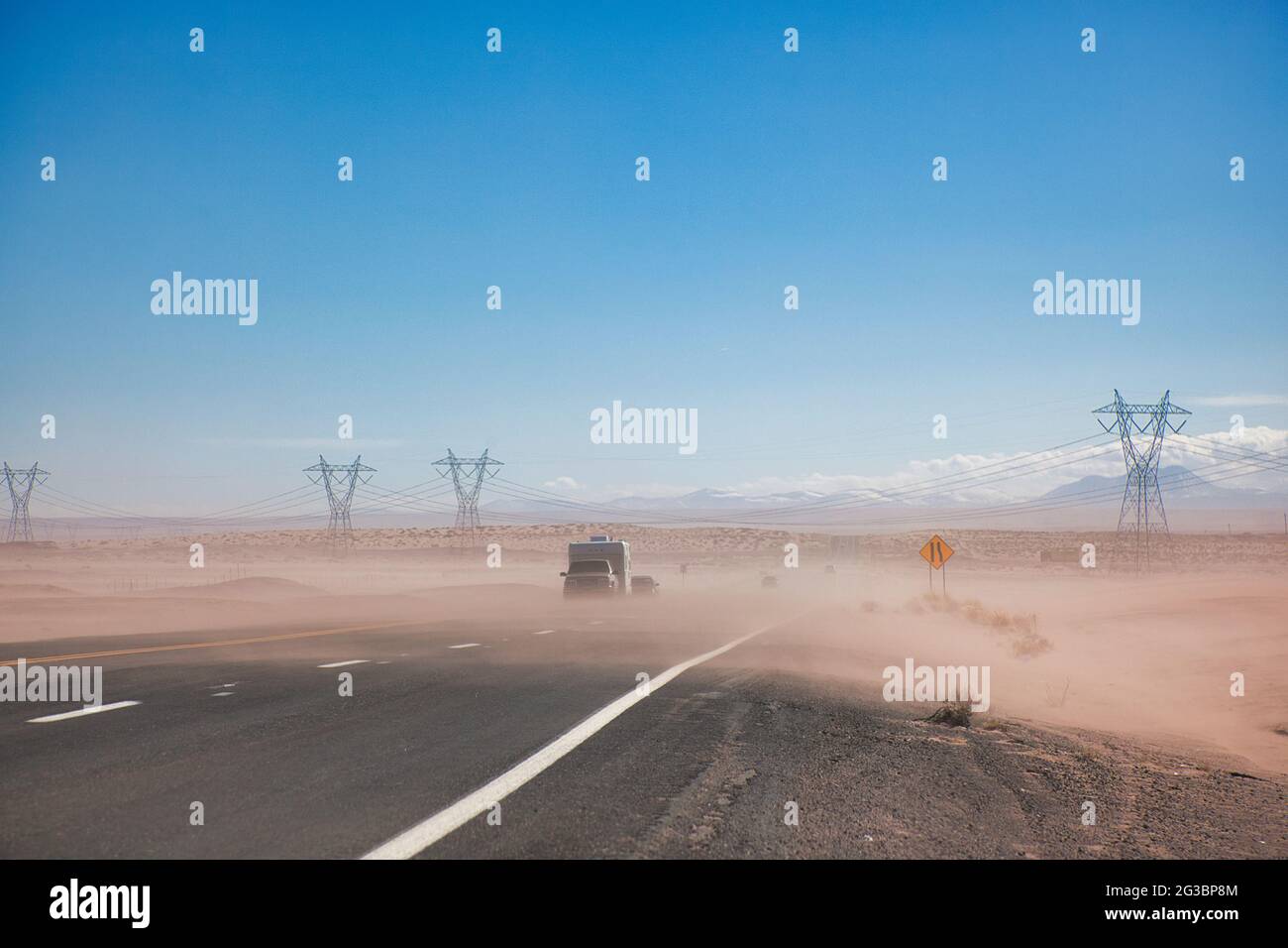 Pilones de electricidad que cruzan una carretera en semi-desierto con una capa de polvo soplando a través de la carretera y dos vehículos que viajan, en Arizona, los Estados Unidos Foto de stock