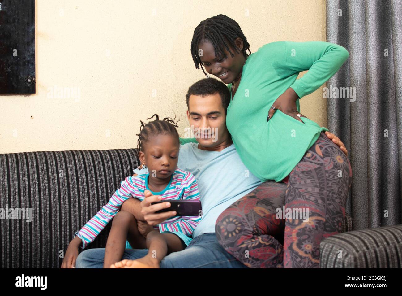 Feliz familia africana mezcla de raza étnica padres con niño pequeño lindo niño hija sentarse en el sofá riendo divertirse sosteniendo el teléfono inteligente usando Foto de stock