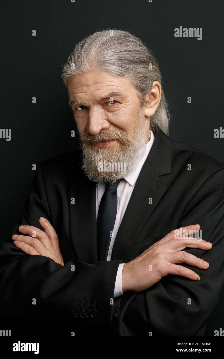 Hombre anciano con estilo que se apuñalaba contra la pared negra con las manos cruzadas. Foto de stock