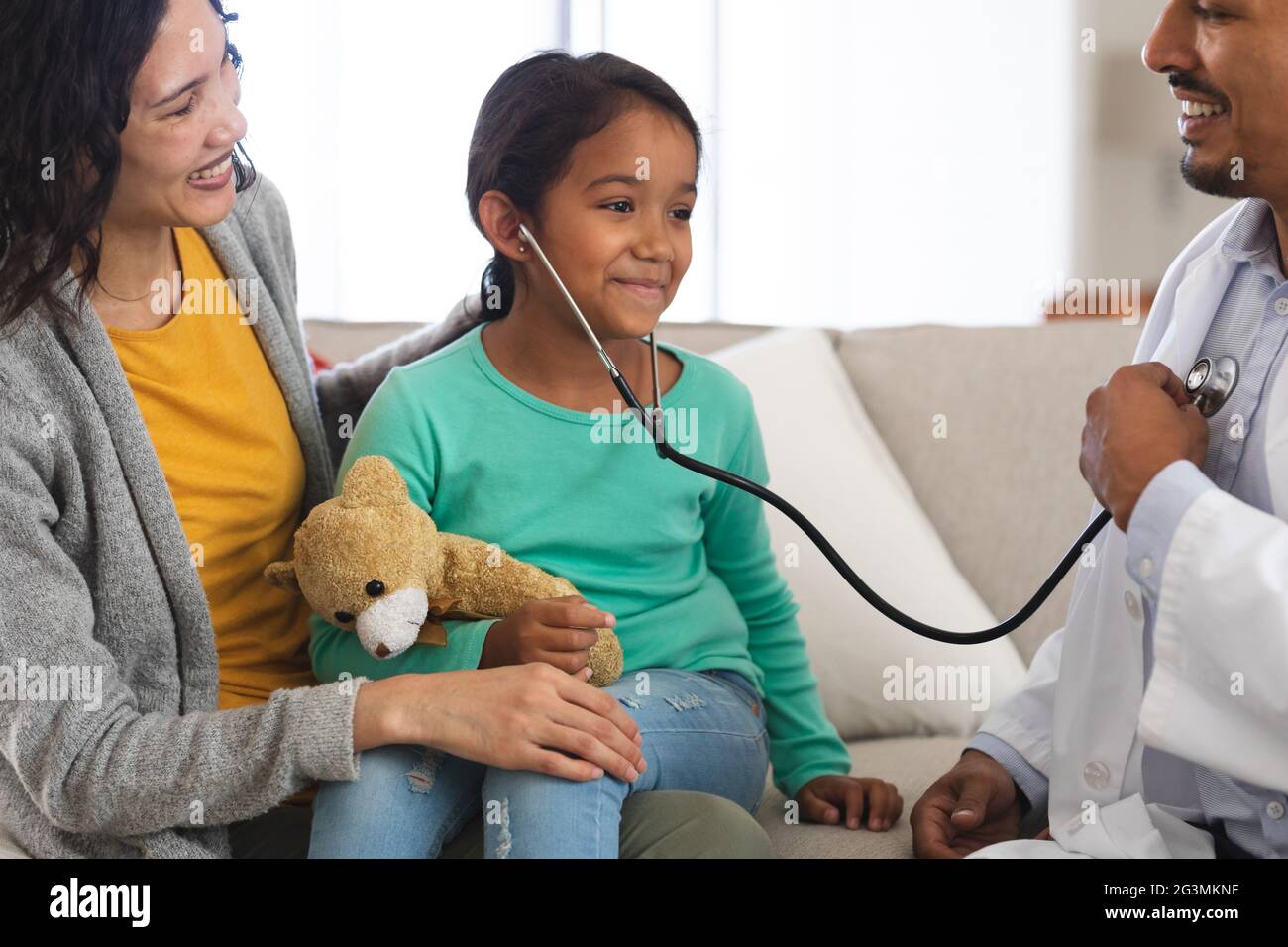 Feliz médico hispano y niña paciente con madre jugando con estetoscopio durante la visita a casa Foto de stock