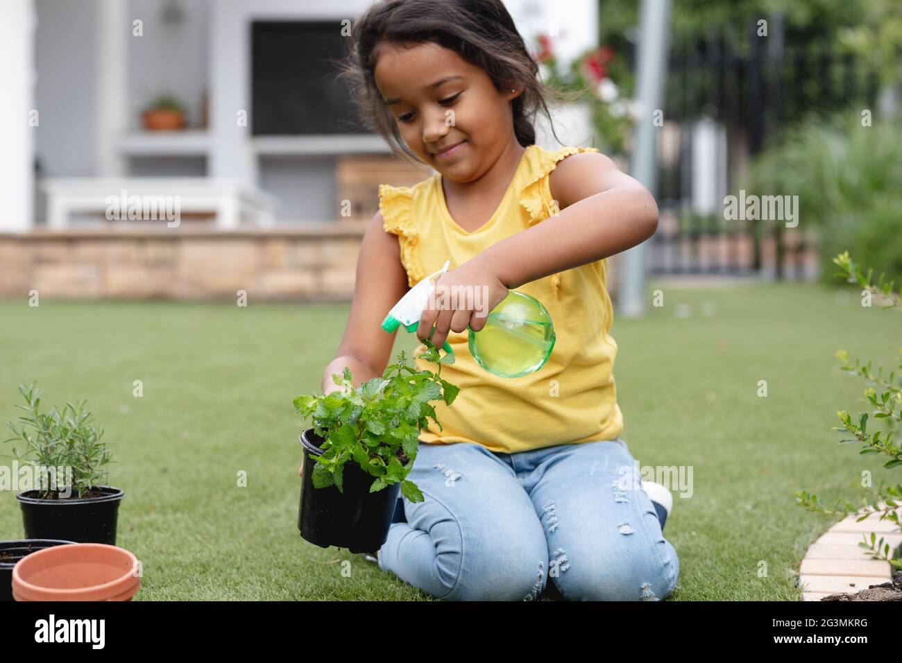 Sonriente niña hispana de jardinería, arrodillarse y riego planta en maceta Foto de stock