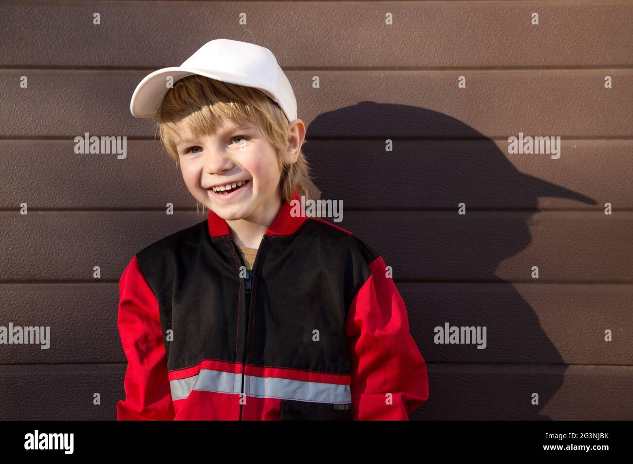 línea letal Actuación Retrato de un hermoso niño de 5 años en un uniforme rojo de un mecánico y  una gorra blanca contra el fondo de una puerta de garaje. Sonríe con un fac  feliz