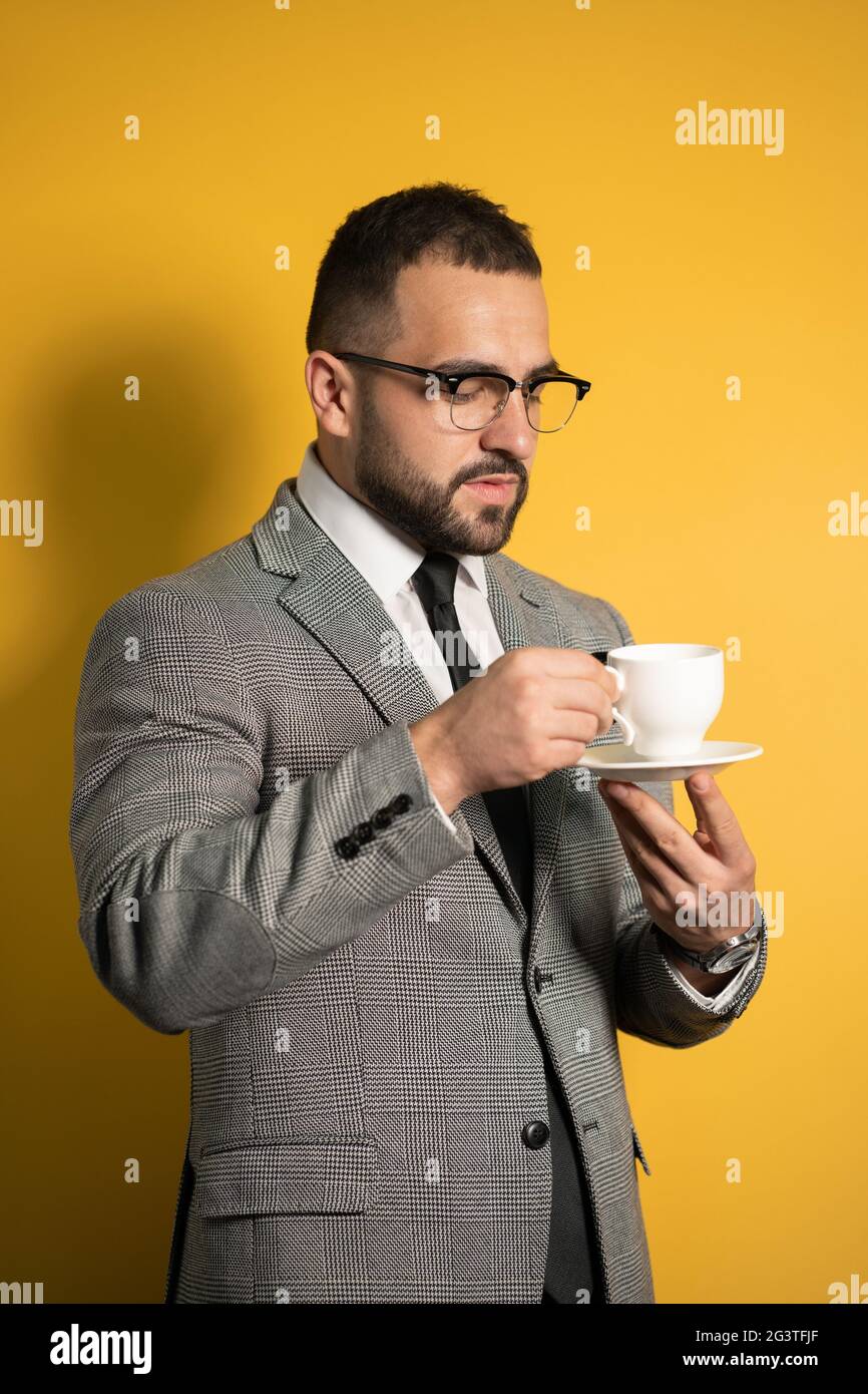Hombre guapo con barba en gafas de ojo en la sujeción formal una taza de café aislada sobre fondo amarillo Foto de stock