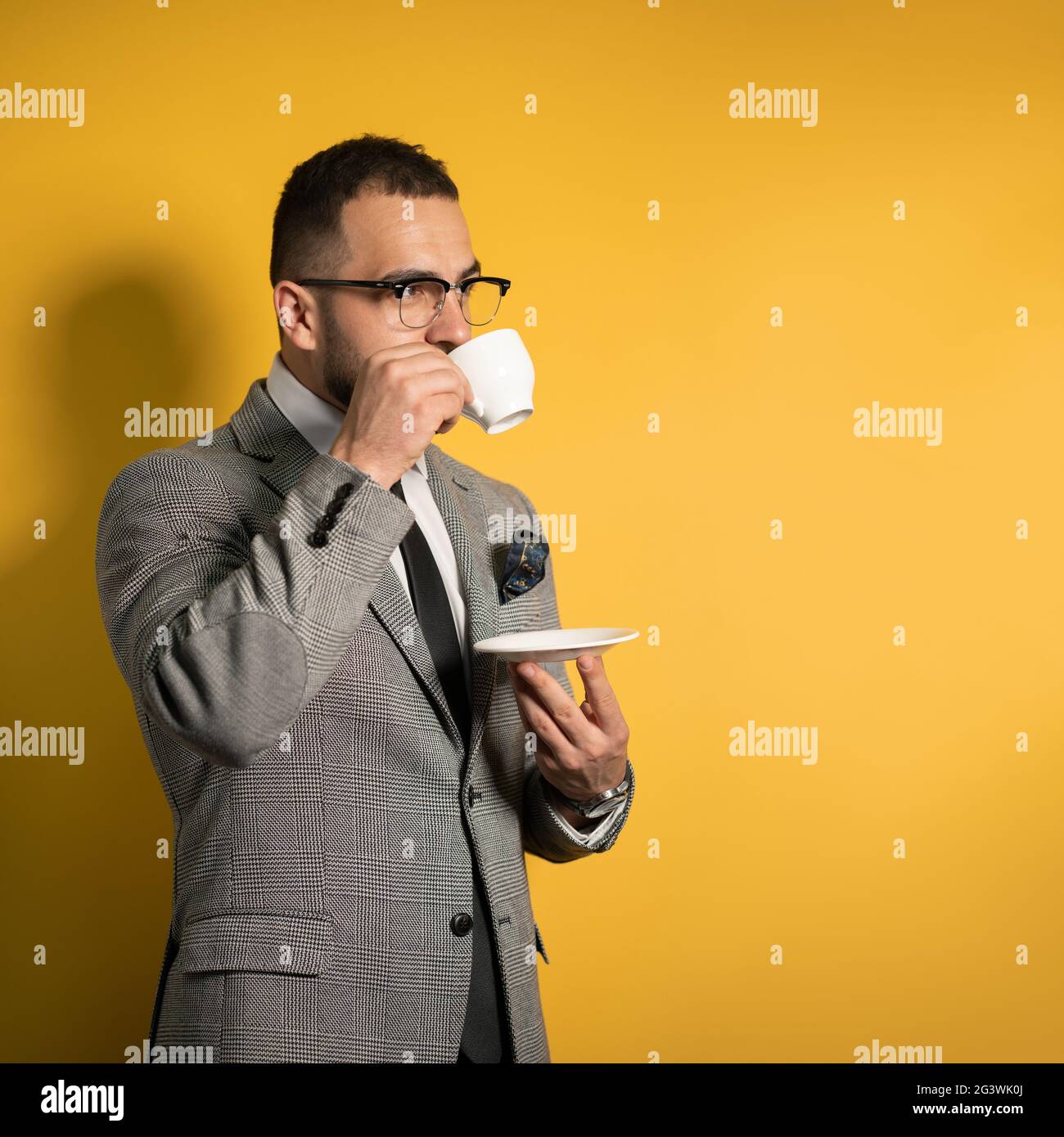 Guapo joven barbudo hombre de negocios en gafas de ojos de vestir con una taza de café de pie aislado lateralmente en amarillo b. Foto de stock