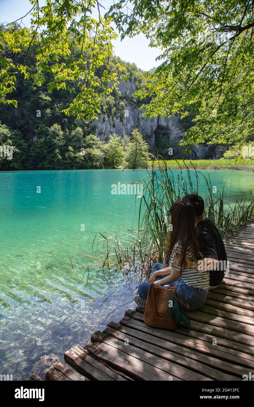 La pareja se sienta en un camino de madera y admira el agua clara en una piscina, el Parque Nacional de los Lagos de Plitvice, Lika-Senj, Croacia, Europa Foto de stock