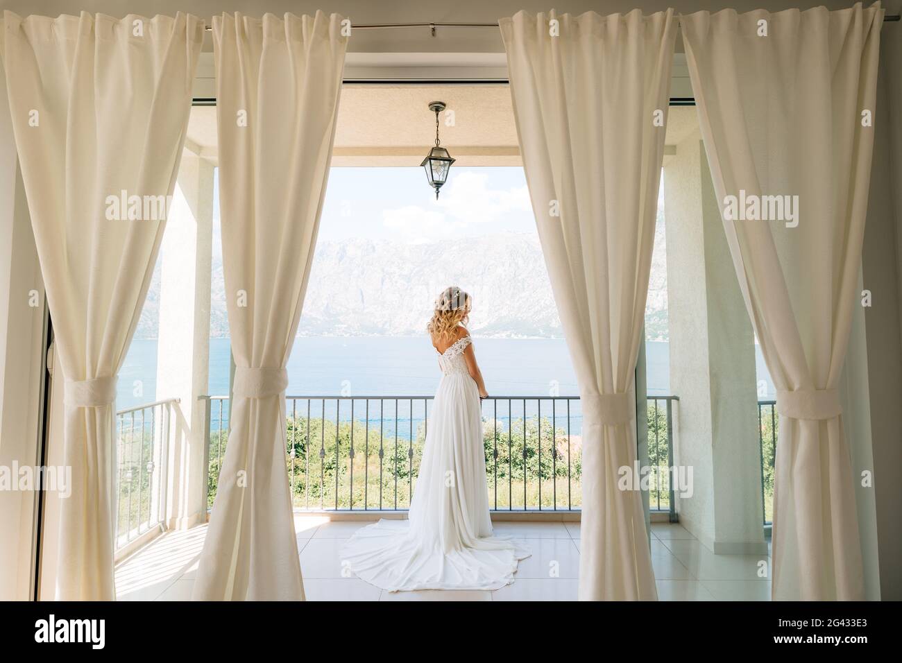 Una novia sofisticada en un elegante vestido de boda se pone de pie un amplio balcón con una vista pintoresca de la bahía Foto de stock