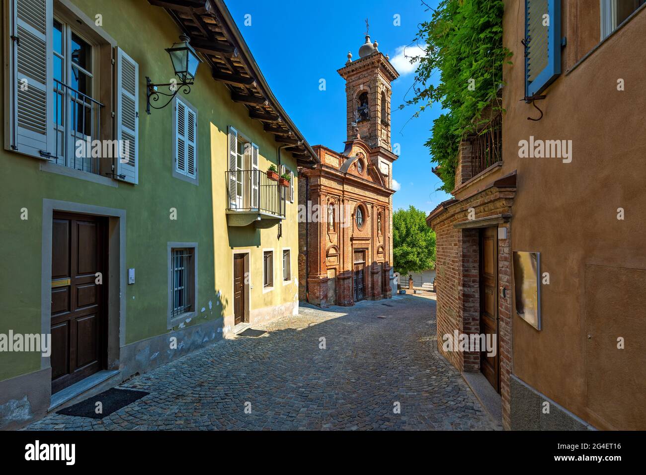 Estrecha calle adoquinada entre casas y antigua iglesia de ladrillo bajo el cielo azul en Piamonte, norte de Italia. Foto de stock
