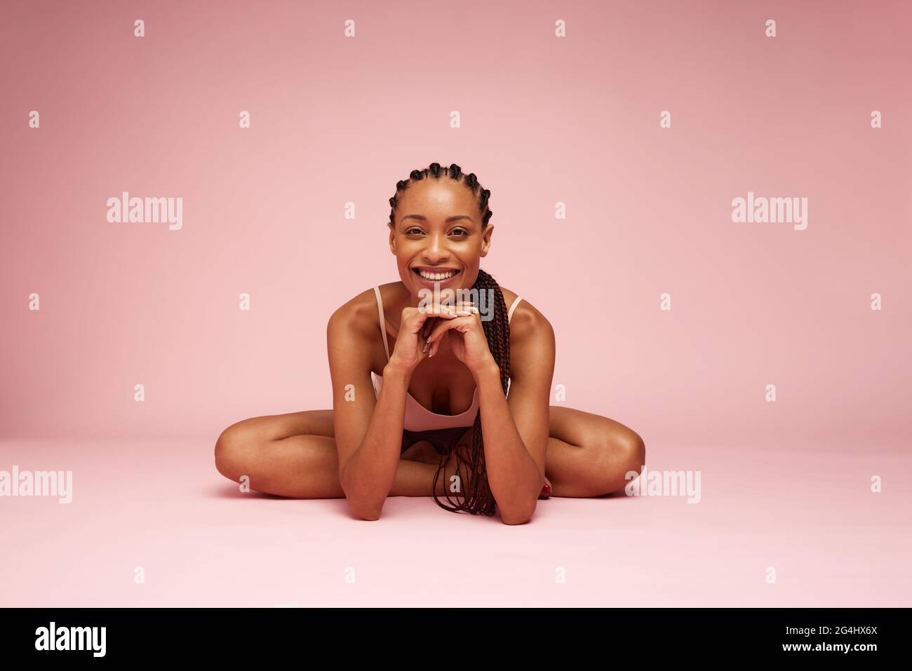Mujer africana bonita sentada en el suelo con la mano en la barbilla. Modelo femenino mirando la cámara y sonriendo mientras está sentado sobre fondo rosa. Foto de stock