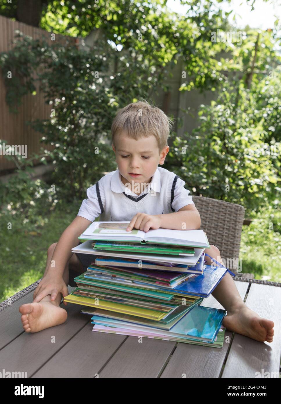 lindo niño se sienta frente a una gran pila de libros para niños sobre un fondo de vegetación en el jardín, bokeh. Libro de lectura enfocado, el develo Foto de stock