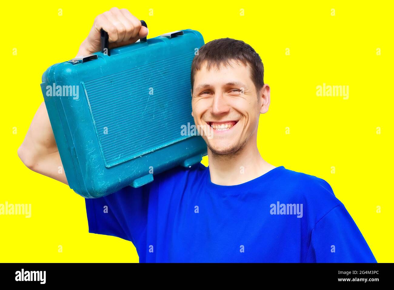 Retrato de un joven reparador positivo sobre un fondo amarillo con una amplia sonrisa. Un trabajador de apariencia caucásica sostiene una caja de herramientas en su hombro. Reparaciones urgentes de guardia. Burlándose Foto de stock