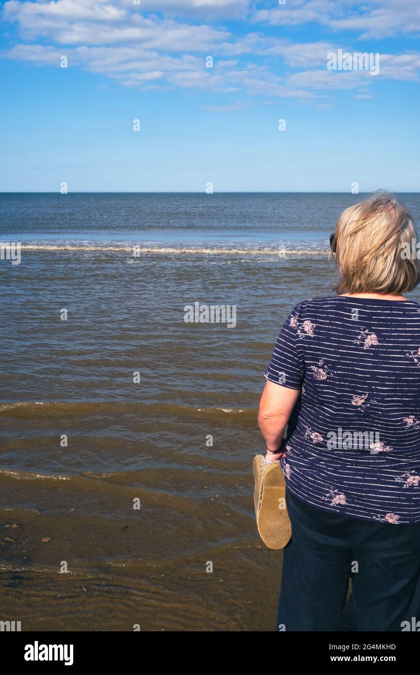 Una mujer rubia disfruta viendo el mar mientras las olas se enrolla en la playa en un hermoso día soleado en el Reino Unido Foto de stock