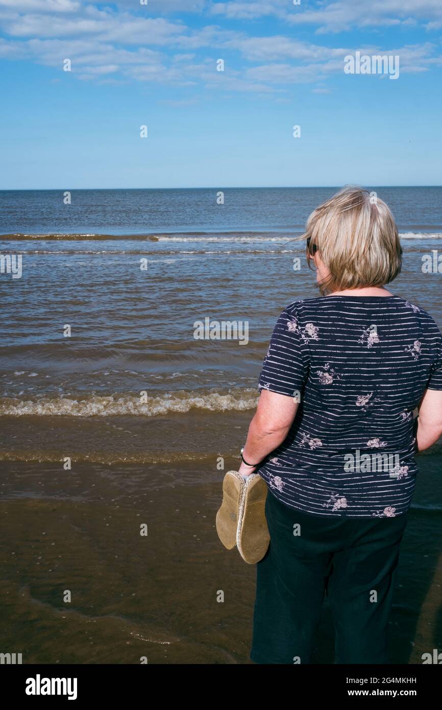 Una mujer rubia disfruta viendo el mar mientras las olas se enrolla en la playa en un hermoso día soleado en el Reino Unido Foto de stock