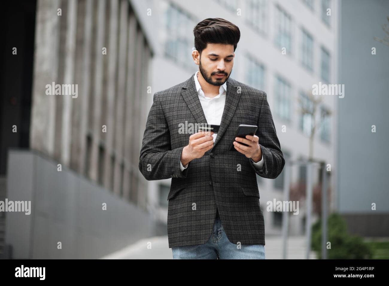 Guapo hindú hombre en ropa formal con estilo con un moderno smartphone y  tarjeta de crédito para compras en línea. Hombre de negocios barbudo de pie  en la calle y haciendo la