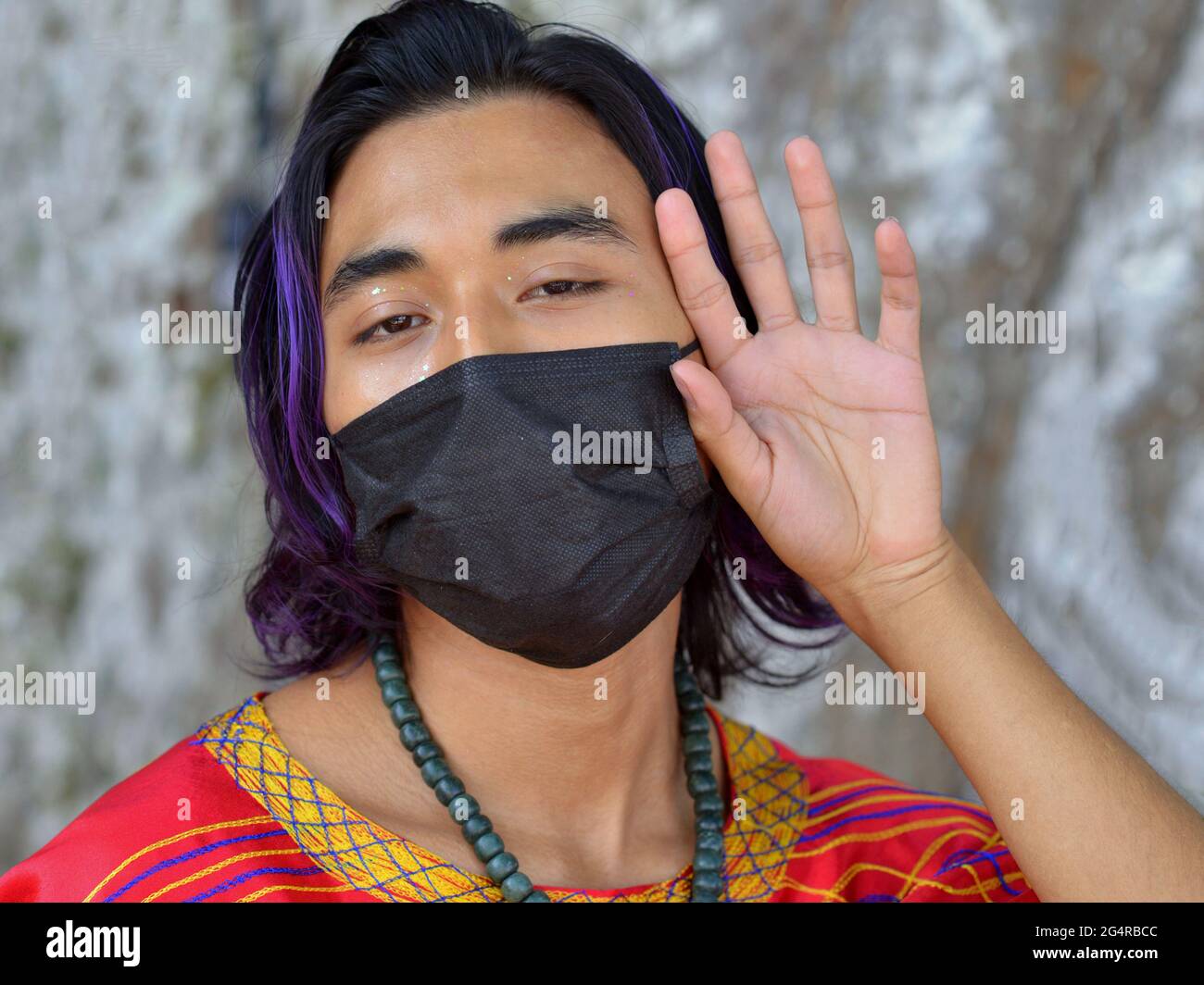 Guapo joven mexicano con azul púrpura teñido pelo streak lleva una máscara de cara negra durante la pandemia mundial de coronavirus y mira la cámara. Foto de stock